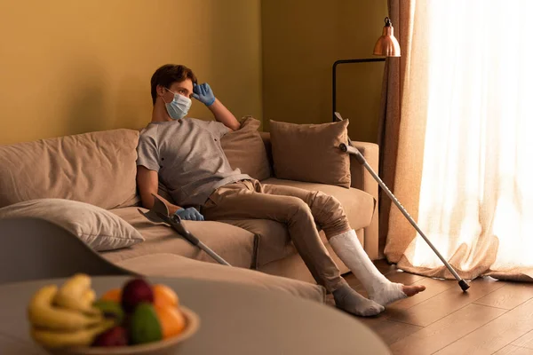 Foco seletivo do homem em máscara médica e bandagem de gesso na perna sentada no sofá em casa — Fotografia de Stock