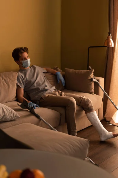 Selective focus of man in medical mask with broken leg sitting on sofa in living room — Stock Photo