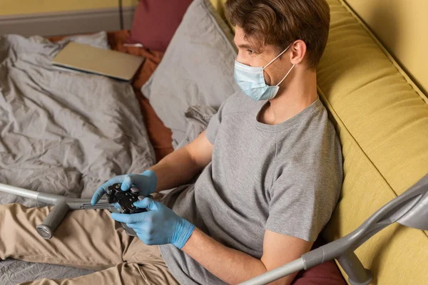 KYIV, UKRAINE - 24 de abril de 2020: Homem de máscara médica e luvas de látex jogando videogame perto de muletas na cama — Fotografia de Stock