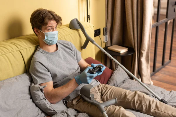 QUIIV, UCRÂNIA - ABRIL 24, 2020: Homem com deficiência em máscara médica e luvas de látex segurando gamepad perto de muletas no quarto — Fotografia de Stock