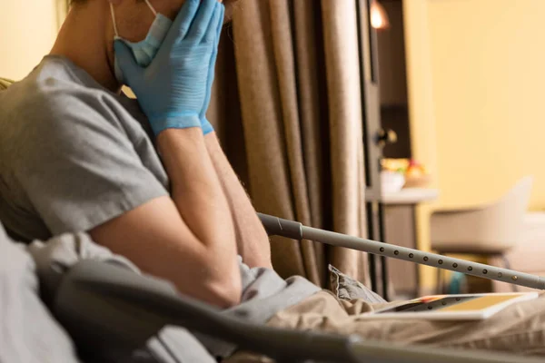 Selective focus of upset man in medical mask and latex gloves covering face near crutches and digital tablet on bed — Stock Photo