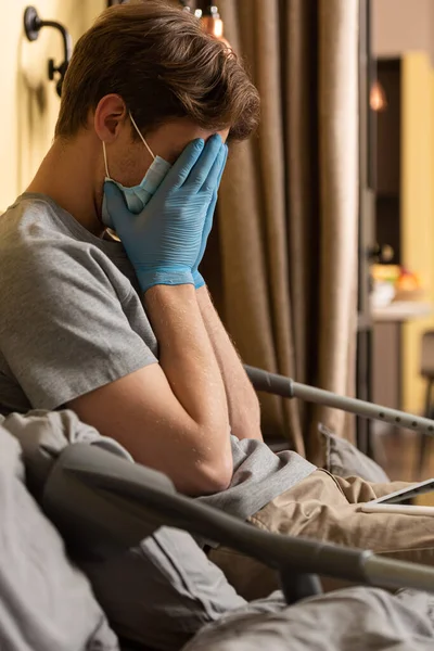 Vista laterale di uomo disabile sconvolto in maschera medica volto di copertura vicino tablet digitale e stampelle sul letto — Foto stock