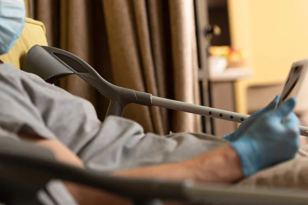 Ausgeschnittene Ansicht eines behinderten Mannes mit medizinischer Maske und Latexhandschuhen, der ein digitales Tablet in der Nähe von Krücken auf dem Bett hält — Stockfoto