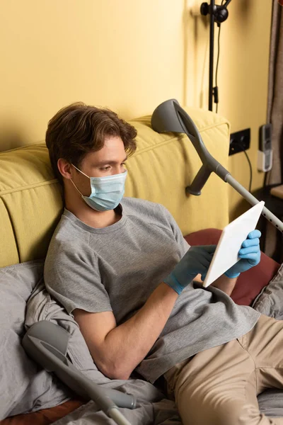 Людина з обмеженими можливостями в медичній масці та латексних рукавичках, використовуючи цифровий планшет біля милиць на ліжку — стокове фото