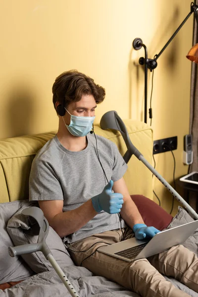 Hombre discapacitado con máscara médica y guantes de látex mostrando como durante la videollamada en el portátil cerca de muletas en la cama - foto de stock