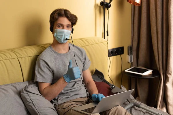 Человек в медицинской маске и наушниках показывает большой палец перед камерой возле костылей на кровати — стоковое фото