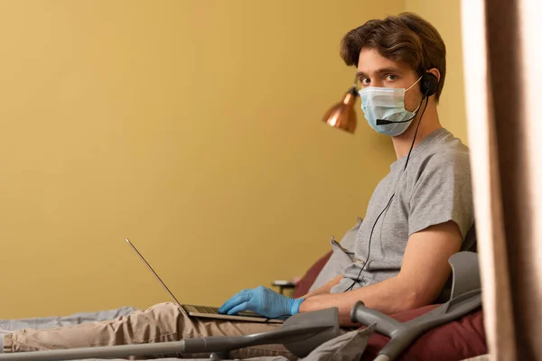 Vista lateral do homem em máscara médica e fone de ouvido olhando para a câmera ao usar laptop perto de muletas no quarto — Fotografia de Stock