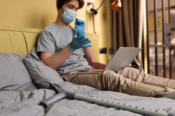 Selective focus of crutch near man in medical mask and headset using laptop during video call on bed — Stock Photo
