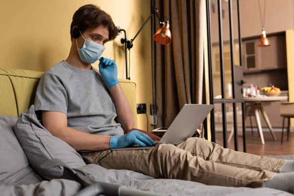 Enfoque selectivo del freelancer en máscara médica con auriculares y portátil cerca de muletas en la cama - foto de stock