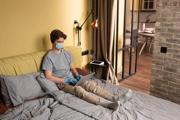 Disabled freelancer in medical mask and latex gloves using headset and laptop near crutches on bed — Stock Photo