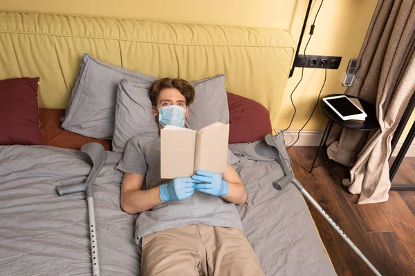 Uomo disabile in maschera medica e guanti in lattice guardando la fotocamera mentre tiene libro vicino stampelle sul letto — Foto stock