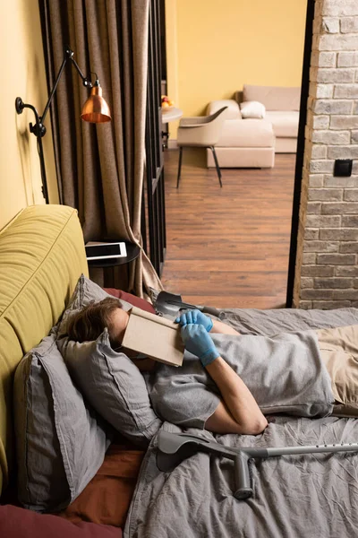 Инвалид в латексных перчатках держит книгу возле лица, когда лежит на кровати рядом с костылями. — стоковое фото