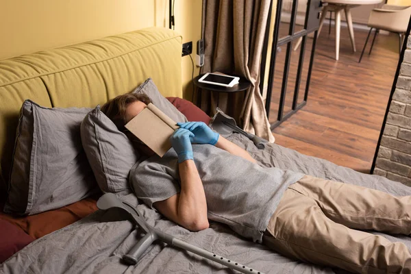 Homem de luvas de látex cobrindo rosto com livro perto de muletas na cama — Fotografia de Stock