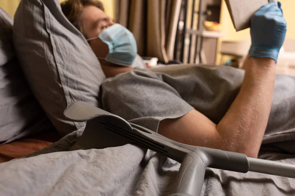 Selektiver Fokus des behinderten Mannes in medizinischer Maske, der Buch in der Nähe der Krücke auf dem Bett liest — Stockfoto