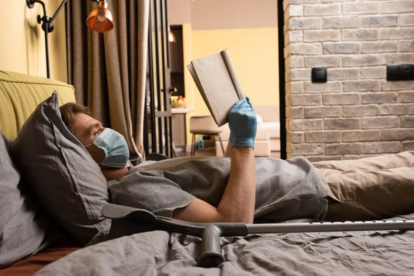 Vista lateral do homem em máscara médica e luvas de látex livro de leitura perto muleta na cama — Fotografia de Stock