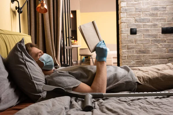 Seitenansicht eines Mannes mit medizinischer Maske und Latex-Handschuhen beim Lesen eines Buches nahe der Krücke auf dem Bett — Stock Photo