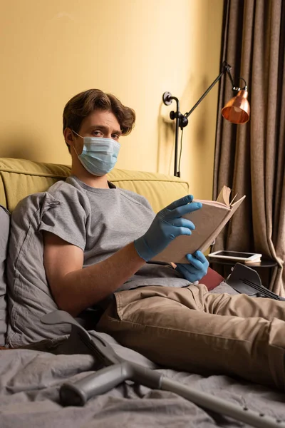 Enfoque selectivo del hombre discapacitado en máscara médica y guantes de látex mirando a la cámara mientras sostiene libro cerca de muletas en la cama - foto de stock