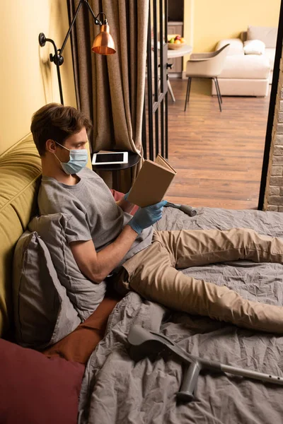 Homem com deficiência em máscara médica livro de leitura perto de muletas na cama — Fotografia de Stock