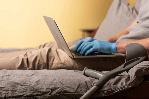 Ausgeschnittene Ansicht einer Krücke in der Nähe eines behinderten Mannes in Latexhandschuhen mit Laptop auf dem Bett — Stockfoto