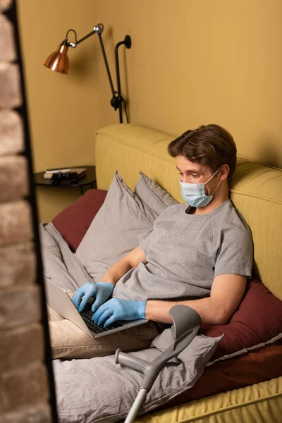 Enfoque selectivo de teletrabajador en máscara médica usando portátil cerca de muleta en el dormitorio - foto de stock