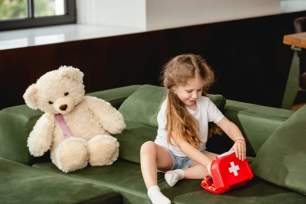 Cute child touching toy first aid kit box near teddy bear while playing doctor game — Stock Photo