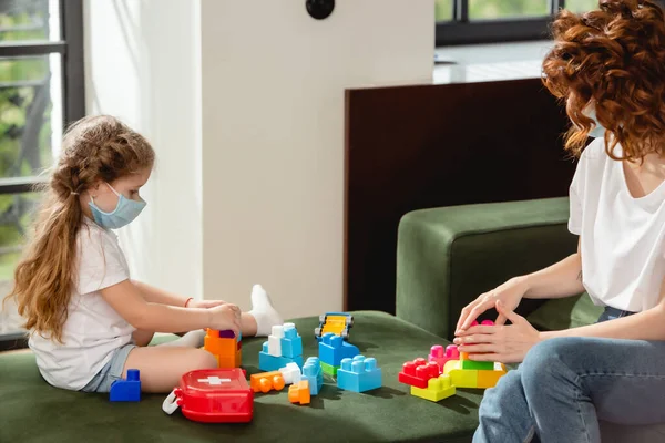 Redhead mother and kid in medical masks playing building blocks — Stock Photo