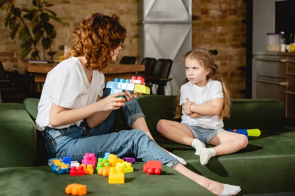 Curly mother holding toy gun and looking at offended daughter with crossed arms — Stock Photo