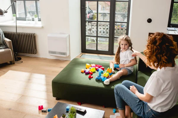 Back view of redhead mother pointing with finger near cute daughter and building blocks on sofa — Stock Photo