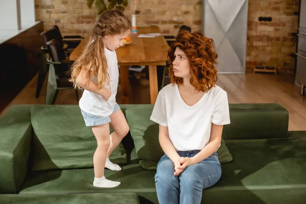 Offended kid standing on sofa and looking at upset and curly mother — Stock Photo