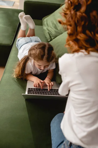 Vista posteriore della madre riccia vicino figlia utilizzando il computer portatile in soggiorno — Foto stock