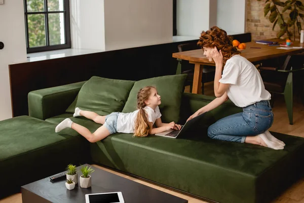 Niedliche Kind mit Laptop in der Nähe lockige Mutter im Wohnzimmer — Stockfoto