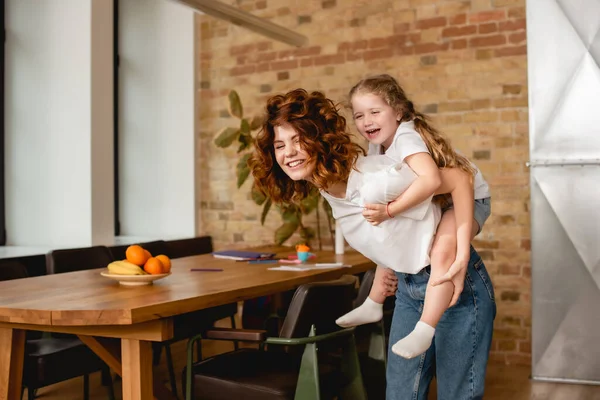 Happy and curly mother piggybacking cute daughter — Stock Photo
