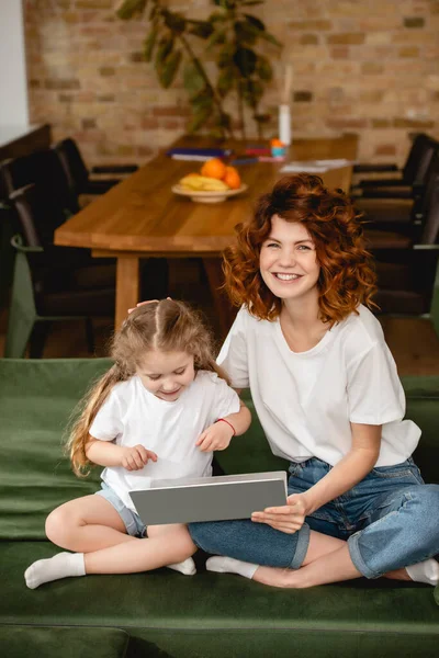 Allegra rossa madre che tiene il computer portatile vicino alla figlia carina in soggiorno — Foto stock