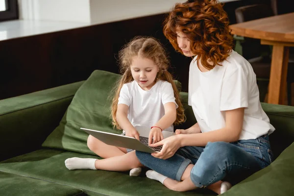 Pelirroja madre sosteniendo portátil cerca adorable hija en sala de estar - foto de stock