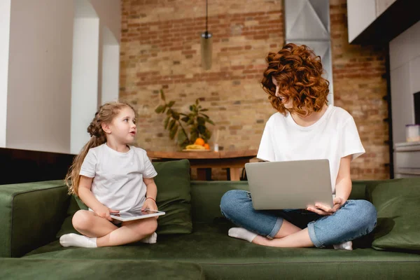 Pelirroja freelancer demandando portátil cerca de adorable hija con tableta digital - foto de stock