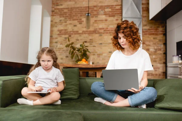 Redhead freelancer utilizzando laptop vicino figlia triste con tablet digitale — Foto stock