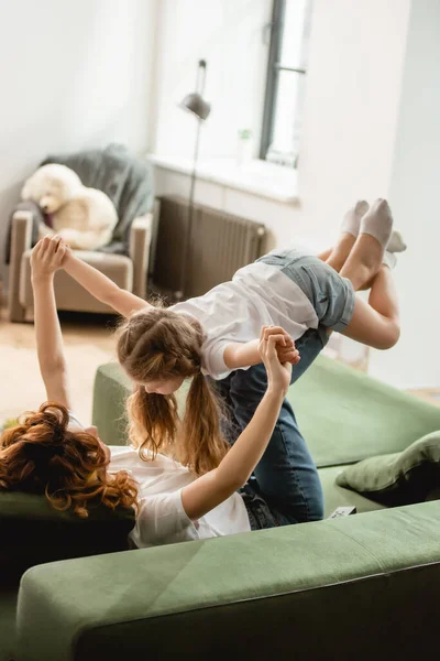 Curly mother lying on sofa, lifting daughter and holding hands in living room — Stock Photo