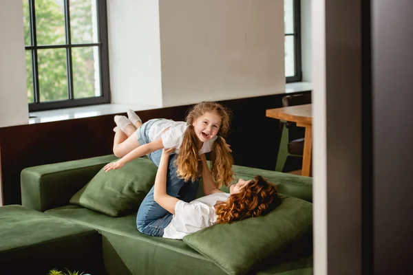 Вибірковий фокус кучерявої матері лежить на дивані, піднімає щасливу дочку і тримає руки у вітальні — стокове фото