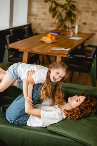 Madre riccia sdraiata sul divano, sollevando figlia allegra e tenendosi per mano in soggiorno — Foto stock