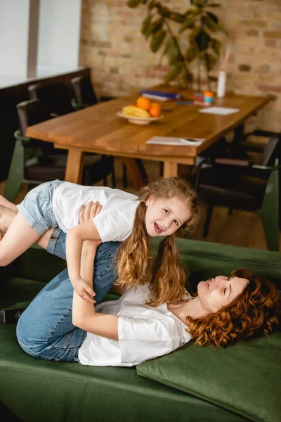 Mère bouclée couchée sur le canapé, soulevant fille gaie et tenant la main dans le salon — Photo de stock