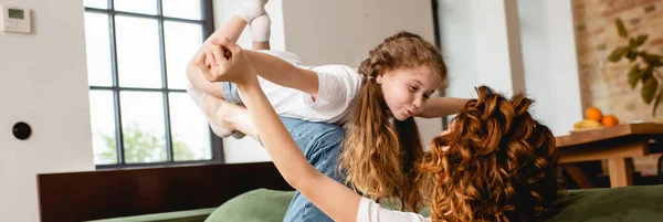 Panoramisches Konzept der lockigen Mutter, die auf dem Sofa liegt, glückliche Tochter hebt und Händchen hält im Wohnzimmer — Stockfoto