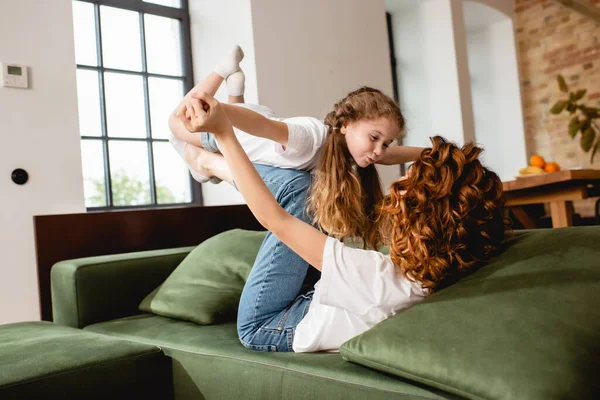 Lockige Mutter auf dem Sofa liegend, niedliches Kind anhebend und Händchen haltend im Wohnzimmer — Stockfoto