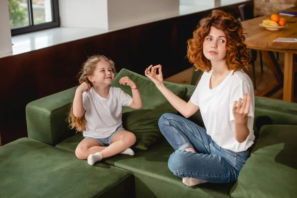 Niño feliz mirando a la madre sentada en postura de yoga en el sofá - foto de stock