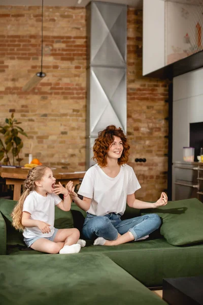 Enfant heureux regardant la mère joyeuse assise dans la pose de yoga sur le canapé — Photo de stock