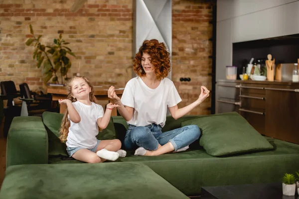 Lockige Mutter sieht Tochter in Lotus-Pose auf Sofa sitzen — Stockfoto