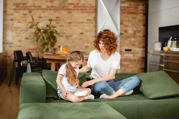 Кудрявая мать смотрит на дочь, сидящую в позе лотоса на диване — стоковое фото