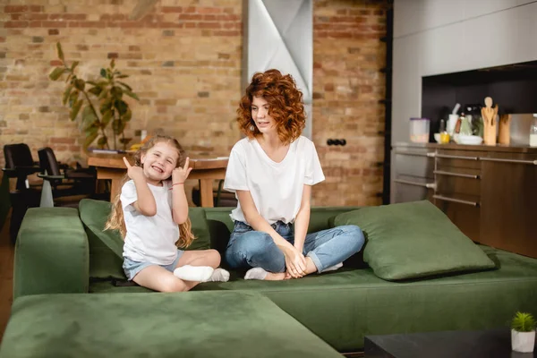 Mère heureuse regardant fille pointant avec les doigts tout en étant assis dans la pose de lotus sur le canapé — Photo de stock