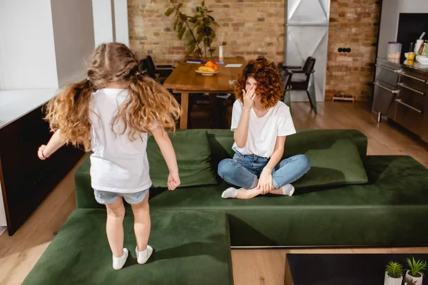 Vue arrière de l'enfant sautant sur le canapé près de la mère frisée couvrant le visage — Photo de stock