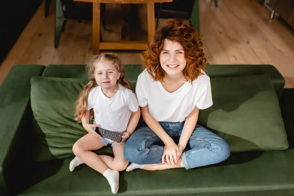 Cute kid holding remote controller and sitting near redhead mother on sofa — Stock Photo