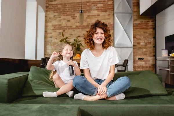 Happy kid holding remote controller and sitting near redhead mother on sofa — Stock Photo